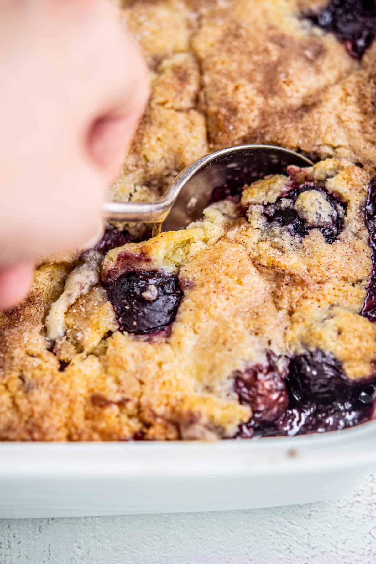 Close-up of a hand using a spoon to serve a portion of a freshly baked cherry and blueberry cobbler in a white dish. The dessert has a golden-brown crust with visible dark berries and a sprinkle of sugar on top.